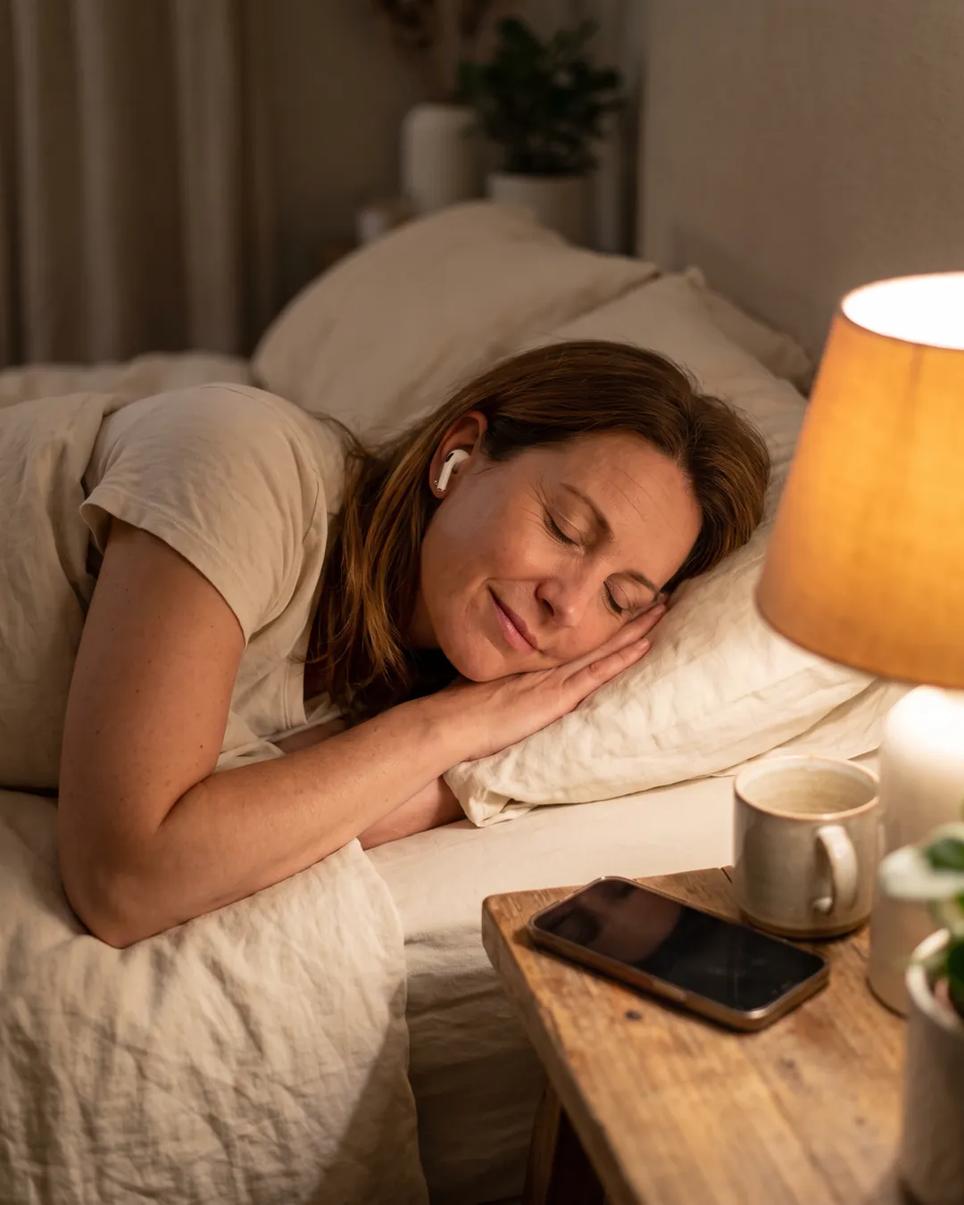 A woman asleep in bed with one earbud in, smiling, under a warm bedside lamp